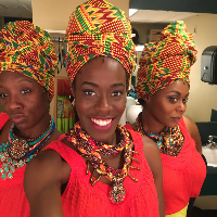 Allison Edwards-Crewe smiling, standing in the middle of two similarly-attired women, all three wearing headwraps and large elaborate woven and beaded necklaces