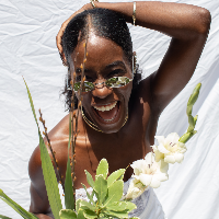 Allison Edwards-Crewe in a lace-like top and sunglasses, laughing and tilted forward, holding the back of her head with her left hand, and white flowers in her right